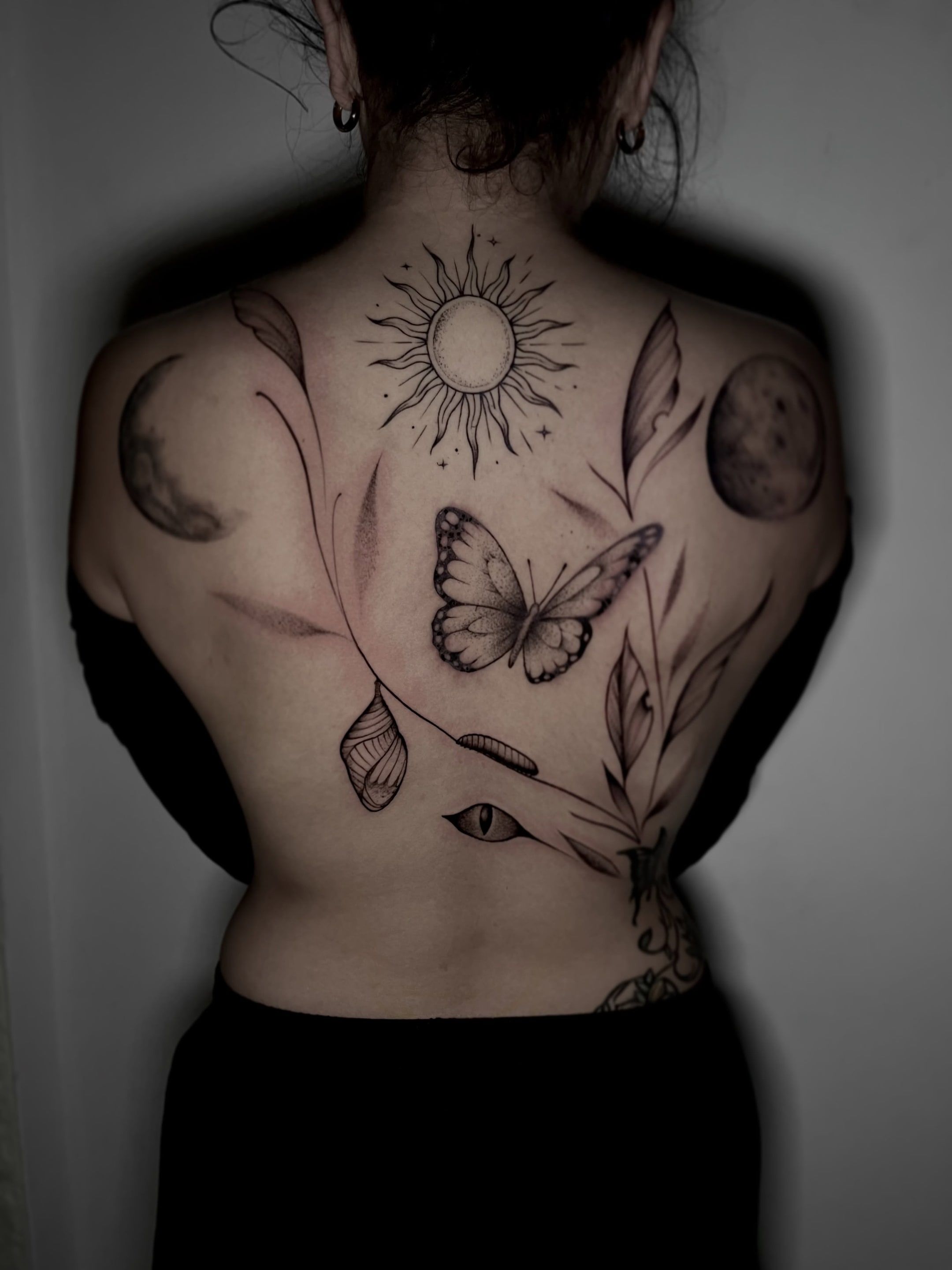 Espalda completa con sol, dos lunas, mariposa, crisálida, oruga, hojas y un ojo en línea fina y sombreado