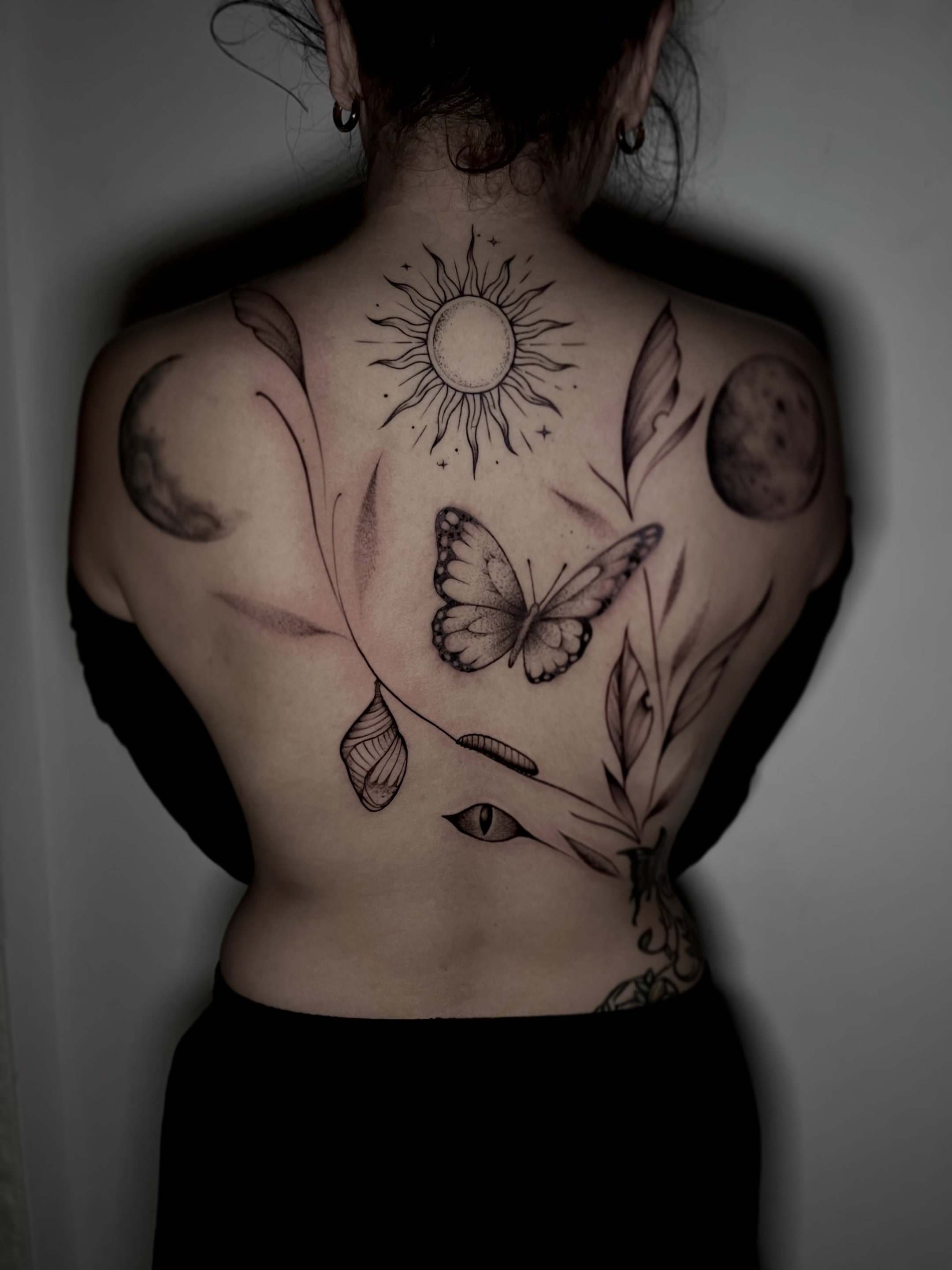 Espalda completa con sol, dos lunas, mariposa, crisálida, oruga, hojas y un ojo en línea fina y sombreado