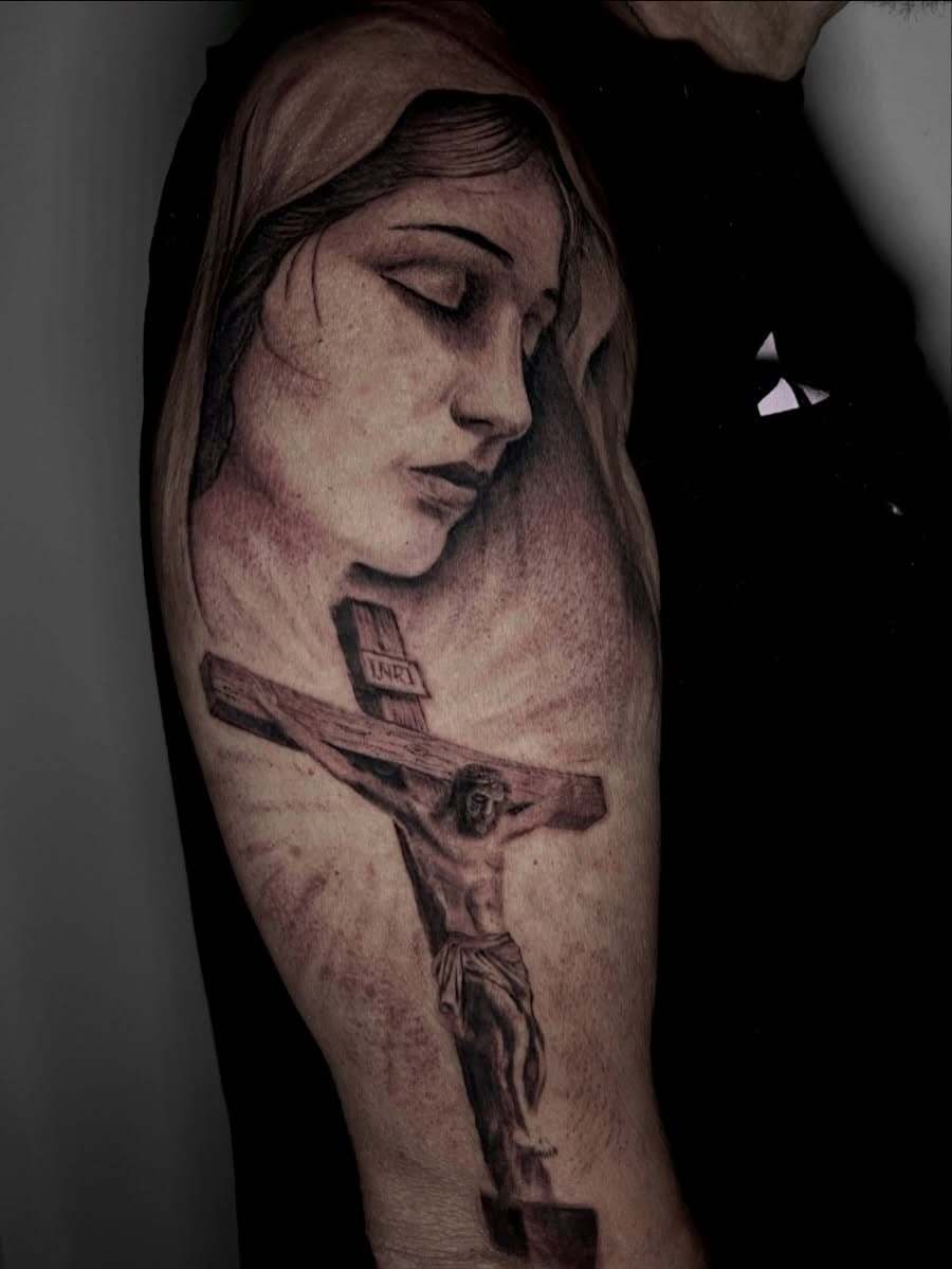 Tatuaje black and grey en el brazo: rostro de la Virgen con los ojos cerrados sobre un Cristo crucificado con rayos de luz